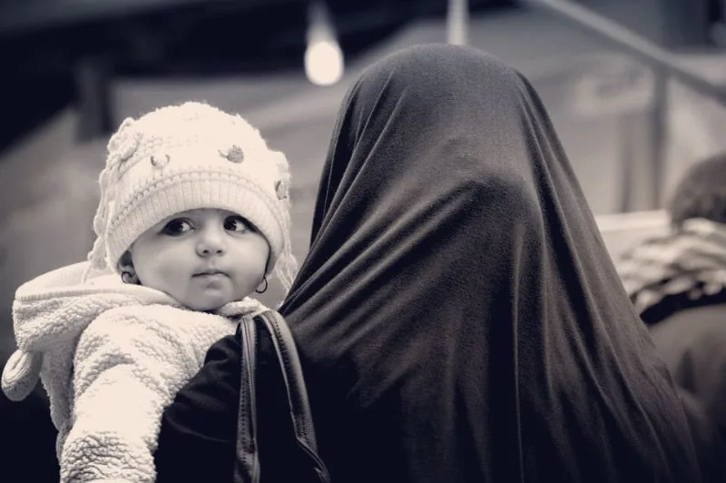Black and white photograph of a baby in a knitted hat held by a veiled mother